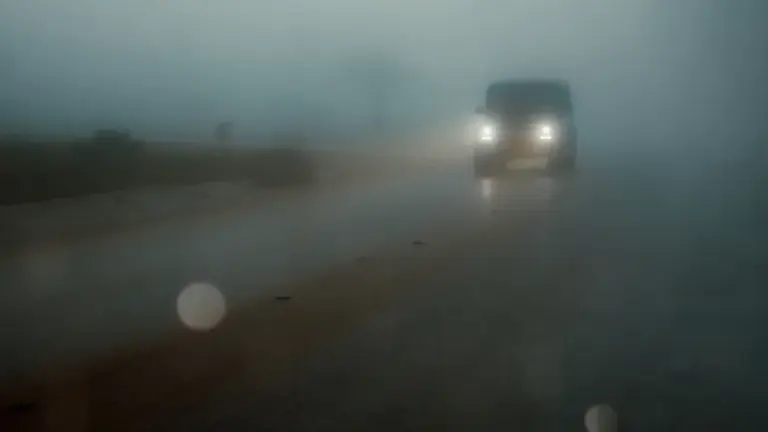 Automašīna brauc pa ceļu biezā miglā ar ieslēgtiem priekšējiem lukturiem sliktos laikapstākļos.