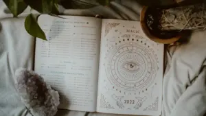 Atvērta dienasgrāmata ar zodiaka zīmju ilustrācijām, kristāliem un augiem uz gultas pārklāja.