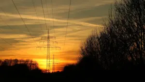 Augstsprieguma elektrības pārvades līnijas saulrietā, simbolizējot elektroenerģijas cenas svārstības Latvijā.