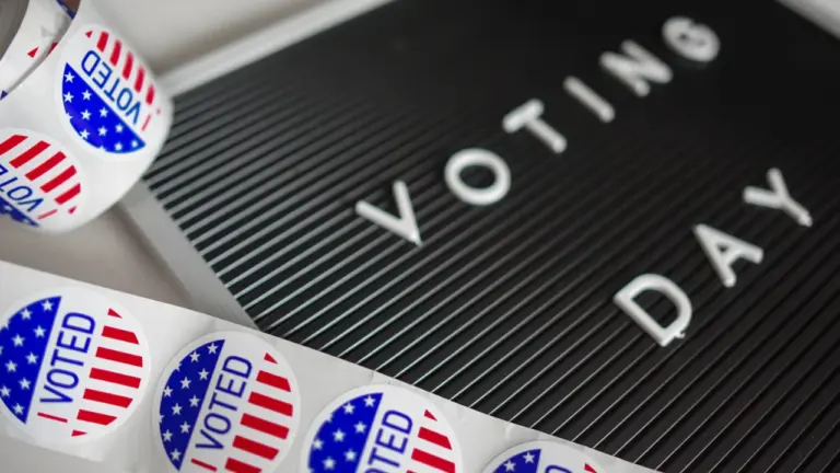 Uzraksts 'Voting Day' uz melna tāfele un balsošanas uzlīmes uz galda.